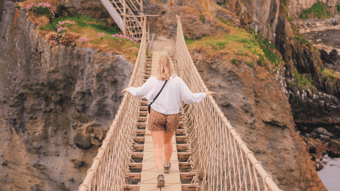 A visitor wearing a grey jumper and leopard print shorts, walking across Carrick-a-Rede Rope Bridge, captured from the island